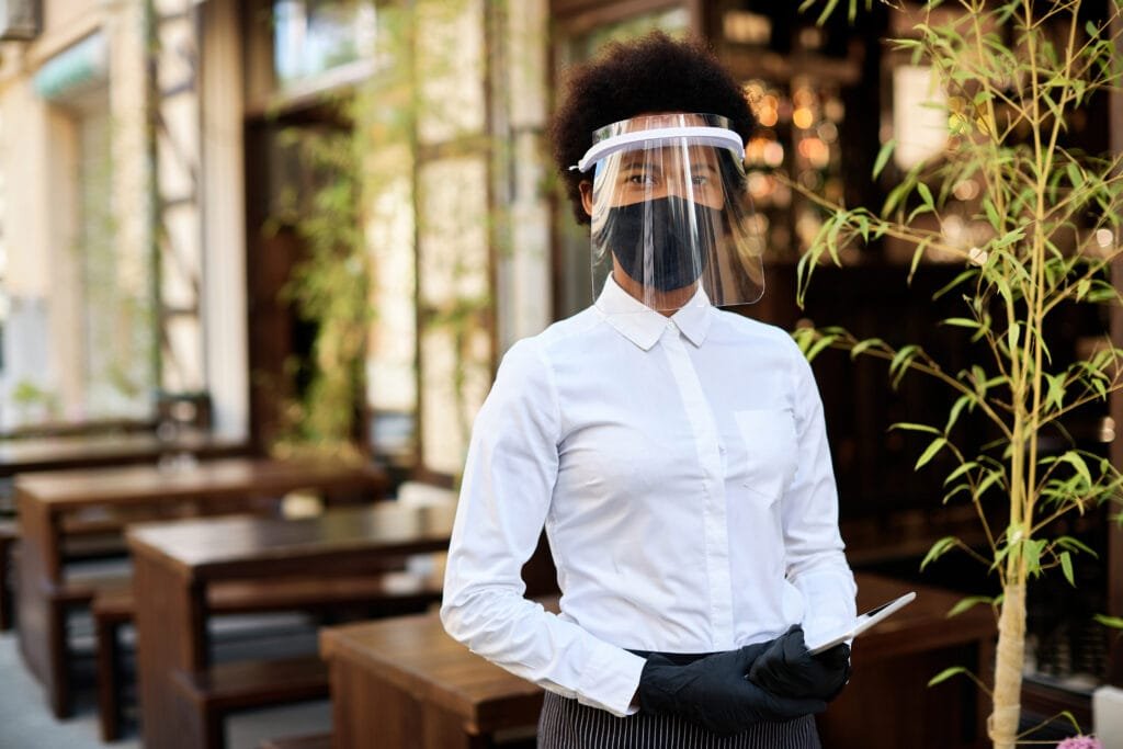 Mesera con careta, cubrebocas y guantes lista para atender en restaurante durante la reapertura segura por COVID-19