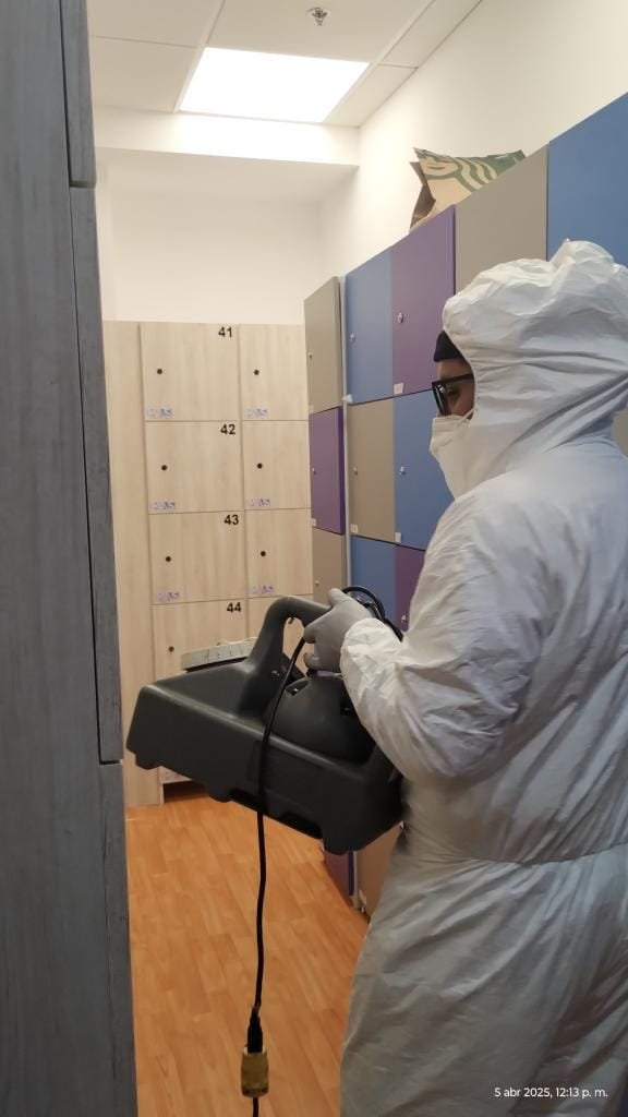 Técnico aplicando fumigación profesional en área de lockers con equipo de nebulización y traje de protección.